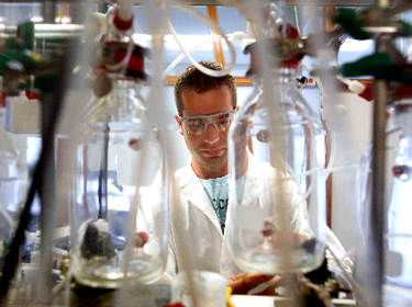 De foto toont een jonge wetenschapper aan het werk in een laboratorium met op de voorgrond glazen flessen en plastic slangen