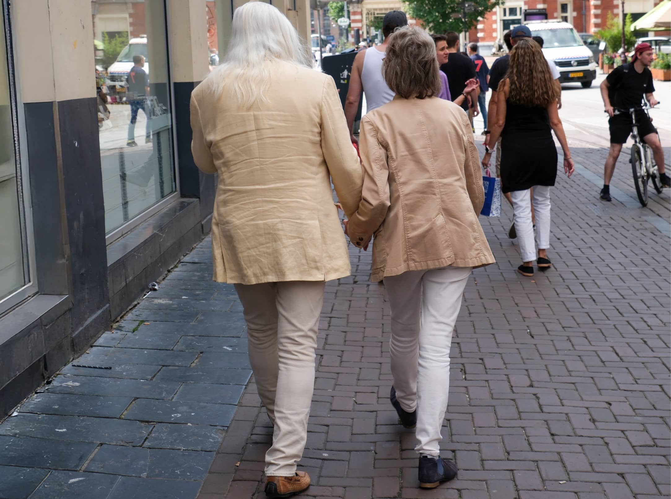 De foto toont een straatbeeld met daarin een koppel dat hand-in-hand wandelt. Ze zijn van de achterkant gefotografeerd.