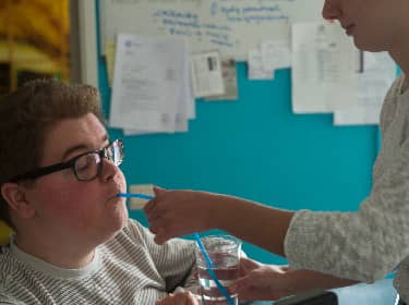 De foto toont een jongen met het Syndroom van Down die water met een rietje krijgt van een verzorgster