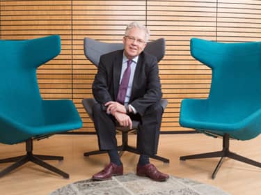 De foto toont Arnold Moerkamp. Hij zit in een grijze stoel, aan beide kanten naast hem staan blauwe stoelen. Moerkamp draagt een zwart pak en een lichtblauw overhemd, donkerbruine schoenen en een paarse das. Hij steunt met zijn ellebogen op zijn bovenbenen en houdt met zijn linkerhand zijn rechterpols vast.