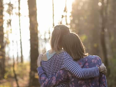 De foto toont twee tienermeisjes met arm om elkaar heen in het bos. We zien de ruggen en de lange haren van de meisjes, de bomen zijn vaag op de achtergrond.