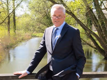 De foto toont Sjaak Wijma. Hij staat op een brug boven een slootje, achter hem zijn het water en bomen te zien. Hij leunt met één hand op de leuning van de brug. Hij draagt een bril en heeft kort grijs haar. Zijn pak is zwart, daaronder draagt hij een lichtblauw overhemd en een grijsblauwe das.