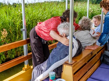 De foto toont een verzorgende die 2 oudere dames een deken geeft tegen de kou. De dames zitten op een bankje buiten, met uitzicht op een veld vol riet bij het water.