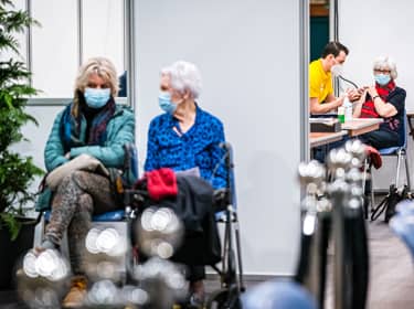 De foto toont twee vrouwen met mondkapjes op in de wachtkamer, met op de achtergrond een arts die een andere vrouw onderzoekt.
