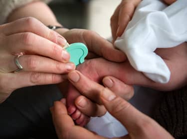 De foto toont een babyvoetje dat door twee mensen wordt vastgehouden, en een hand die met een apparaatje een prik in de hiel zet.