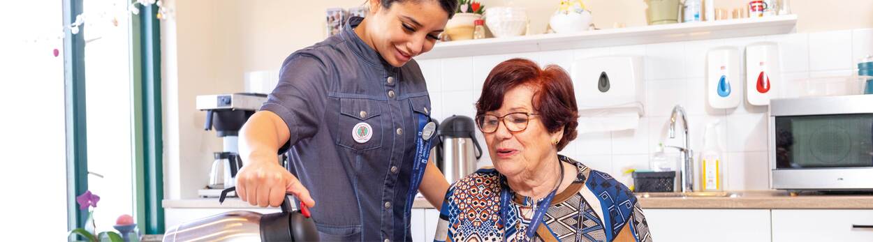 Op de foto zit een oudere vrouw in haar keuken. Rechts van haar staat een thuiszorgmedewerkster die thee inschenkt.