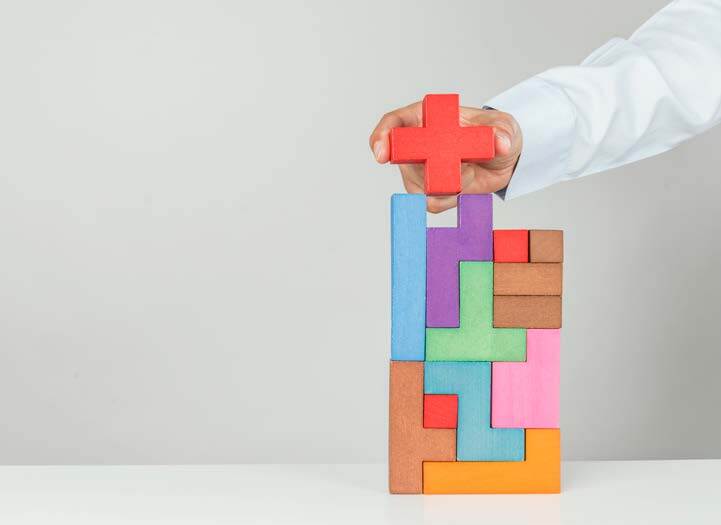 Op de afbeelding is een stapel blokjes te zien. De blokjes hebben vormen die precies in elkaar passen. Een hand voegt een blokje toe in de vorm van een rood kruis. Het blokje past precies in de stapel.