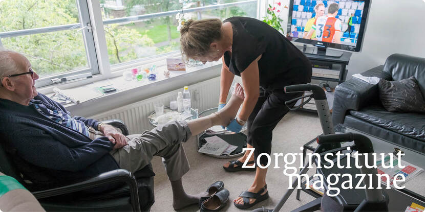 Een oudere man zit in een stoel. Een vrouw bukt zich om hem een steunkous aan te trekken. Naast de man staat een rollator.