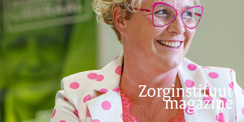 Je ziet Wieke Paulusma, een vrouw met kort blond haar en roze bril in een wit met roze gestipt pak die lacht