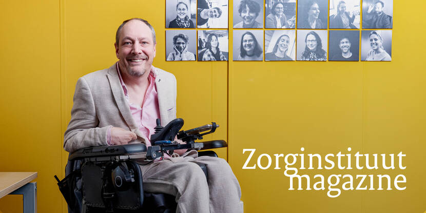 Je ziet Sander Hilberink lachend in pak in een rolstoel met foto's van mensen aan de muur op de achtergrond