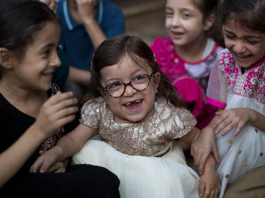 De foto toont een lachend klein meisje met bril in een feestelijke jurk die voortanden mist te midden van andere lachende kinderen