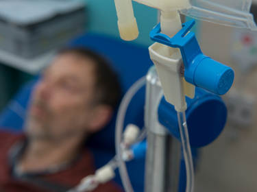 De afbeelding toont een close-up van een infuus en op de achtergrond een vermagerde patiënt