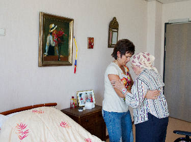 De foto toont een verpleegster die een oudere vrouw met hoofddoek helpt om op te staan in haar slaapkamer
