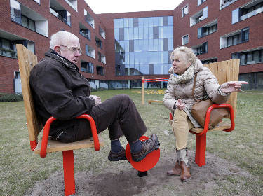 De foto toont een verpleegster die tegen een oudere man praat op twee stoelen in het grasveld voor het moderne verpleegtehuis
