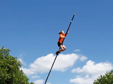 De foto toont een tienermeisje met een polsstok die gaat hoogspringen met op de achtergrond een blauwe lucht