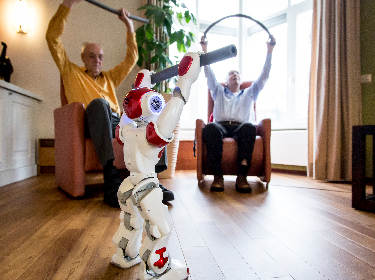 De foto toont een huiskamer waarin twee oudere mensen in leunstoelen een robot nadoen die een foam stok omhoog houdt