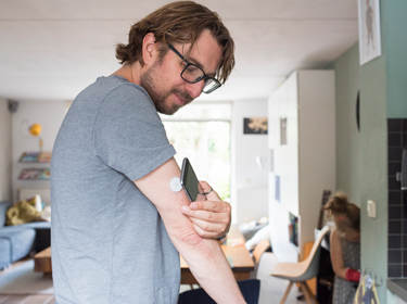 De foto toont een diabetespatiënt die gebruikmaakt van een Flash Glucose Meter. Hij scant het witte apparaatje op zijn bovenarm met zijn smartphone.