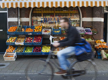 De foto toont iemand die voorbij fietst, met op de achtergrond een groentekraam