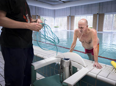 De foto toont een man in een zwembad die oefentherapie volgt voor COPD. De man klimt net uit het zwembad. Voor hem staat iemand met zwarte kleren die de zuurstofdraad vasthoudt die naar de neus van de man loopt. De draad is verbonden aan een zuurstoftank die op een stoel staat tussen deze twee personen in.