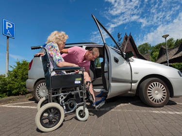 De foto toont een vrouw in een rolstoel die in een auto wordt geholpen