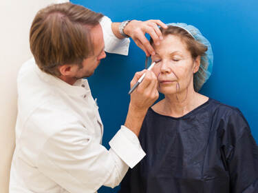 De foto toont een arts en een patiënt voorafgaand aan een cosmetische ingreep. De arts tekent lijnen op het gezicht van de patiënt.