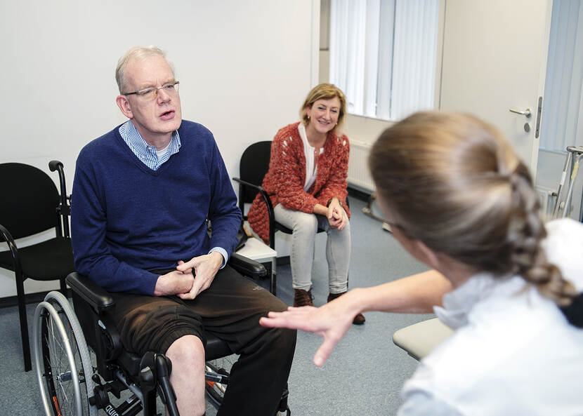 De foto toont een wat oudere man in een rolstoel die luistert naar de uitleg van een arts. Naast de man zit een vrouw die meeluistert en lacht.
