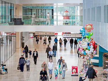 De foto toont de hal van een ziekenhuis. Er lopen verschillende mensen door de hal en rechts is een stand met ballonnen.