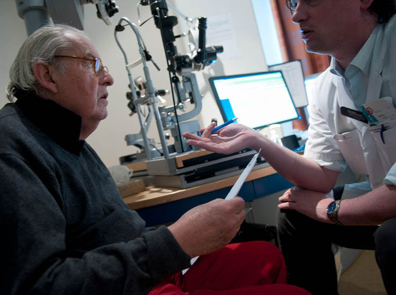 De foto toont een wat oudere man in gesprek met een verpleegster met op de achtergrond computers en een medisch apparaat.