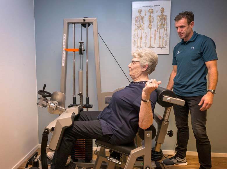 De afbeelding toont een oudere dame die oefent op een trainingsapparaat met gewichten terwijl de mannelijke fysiotherapeut achter haar staat en meekijkt.