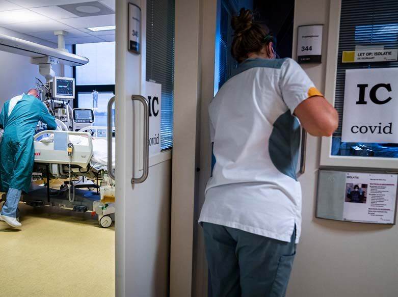 De foto toont een gang van een ziekenhuis. Door een openstaande deur is een kamer te zien waar een verpleger in blauwe beschermende kleding over een ziekenhuisbed gebogen staat. In de gang zien we een verpleegster op de rug. Zij draagt witte kleding. Aan de deur en een raam hangen bordjes met de tekst 'IC COVID'.