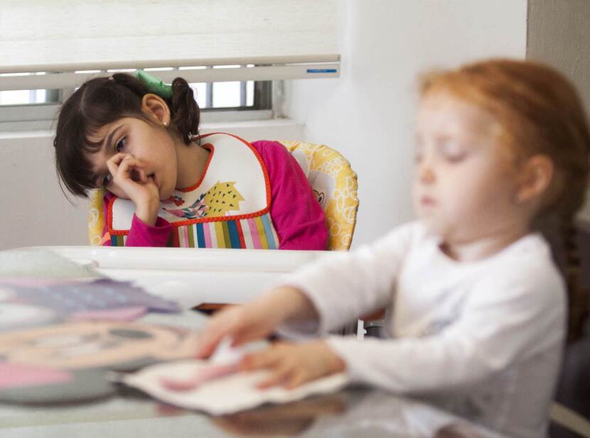 De foto toont twee jonge kinderen die aan een tafel zitten. De foto stelt scherp op een meisje met donker haar dat haar duim in haar mond heeft en rechts opzij in slaap lijkt te vallen. Een meisje met rood haar in de voorgrond speelt met wat papieren.