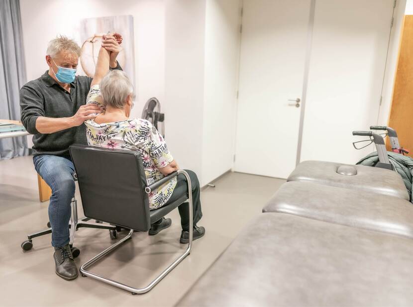 De foto toont een oudere vrouw bij de fysiotherapeut. De vrouw zit in een stoel in een behandelkamer, we zien haar rug. De fysiotherapeut tilt de linkerarm van de vrouw omhoog.