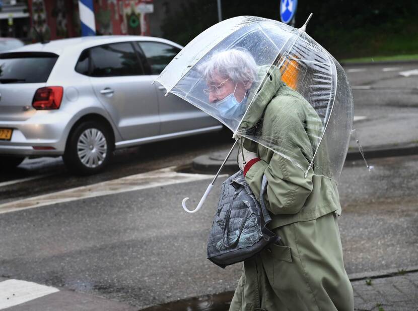 De foto toont een oudere dame op straat. Ze houdt een doorzichtige paraplu vast en draagt een mondkapje over haar mond, onder haar neus. Op de achtergrond is een grijze auto te zien.