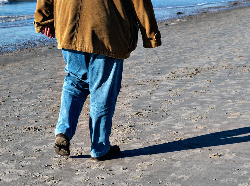 De afbeelding toont de achterkant van een man die wandelt op het strand, waarbij alleen zijn benen en een deel van zijn rug zichtbaar zijn. De man is gekleed in een spijkerbroek en lichtbruine jas.