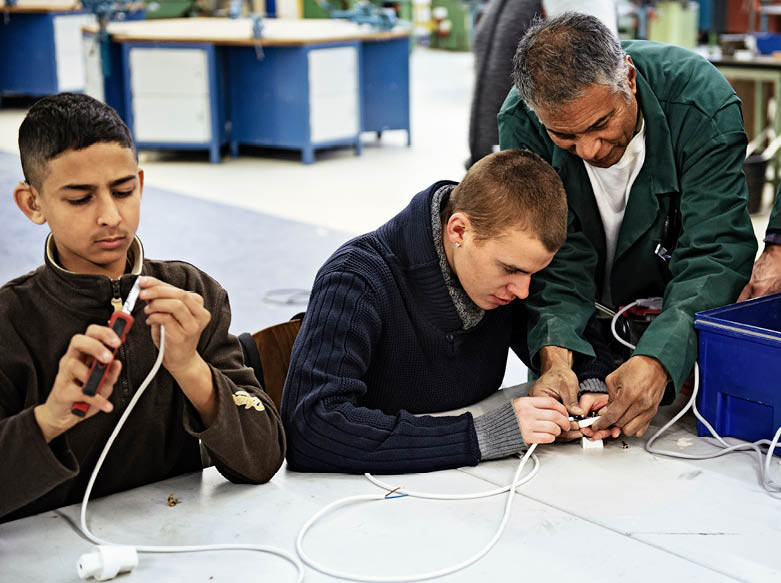 De afbeelding toont twee jongens die aan een tafel zitten en van de naast hen staande beleider uitleg krijgen over hoe ze een stekker aan een kabel moeten monteren.