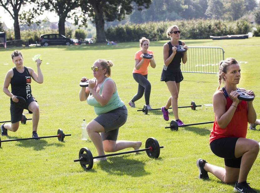 De foto toont 5 mensen die in een park aan het sporten zijn. Ze houden een gewicht vast en doen lunges. In het gras liggen halters met gewichten eraan.