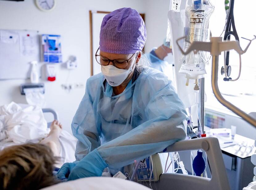 De foto toont een zorgverlener bij een patiënt in een ziekenhuisbed. De foto is van achter het ziekenhuisbed gemaakt, we zien alleen de bovenkant van het hoofd en een been van de patiënt. De zorgverlener draagt blauwe beschermende kleding, een paars haarnetje en een mondkapje en buigt zich over de patiënt. Naast het bed hangt een infuus.