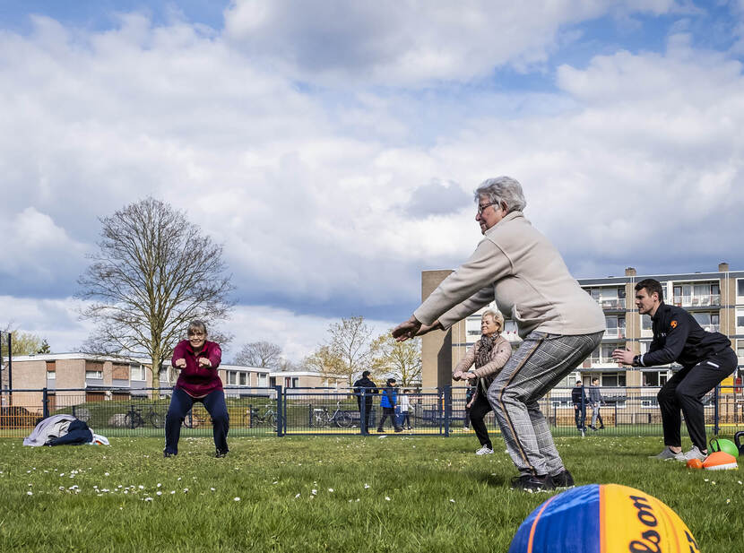 De foto toont drie oudere vrouwen die onder leiding van een jongere man sporten in het park.