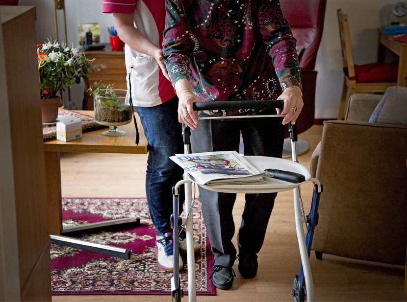 De foto toont een oudere mevrouw die met een rollator door een woonkamer loopt, daarbij geholpen door een zorgverlener die achter haar staat. Alleen hun lichamen zijn in beeld, niet hun gezicht.
