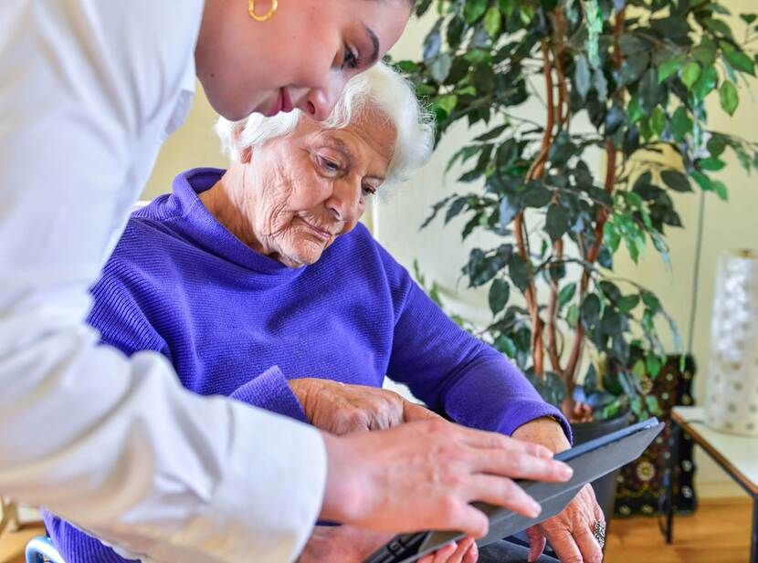 De foto toont een oudere vrouw op een stoel met een paarse trui aan. Voor haar staat een jongere vrouw gebogen en samen kijken ze naar het scherm van een tablet.