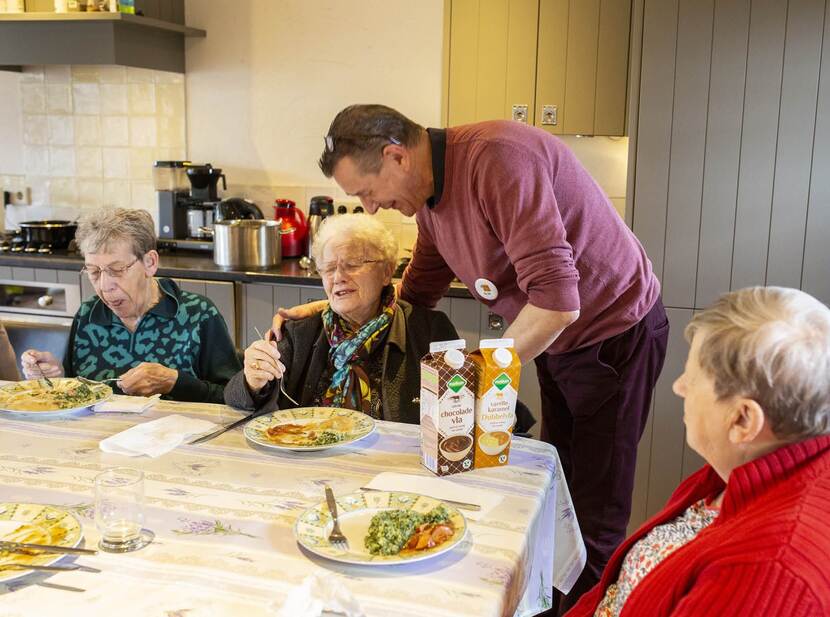 De foto toont een groep ouderen die eten aan een tafel en een verzorger die 1 van de personen helpt.