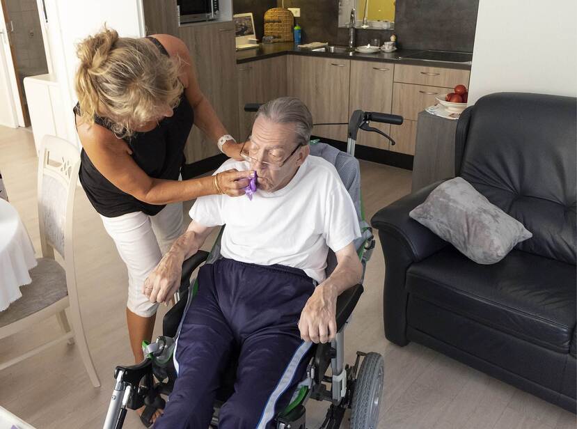De foto toont een oudere man in een rolstoel. Hij wordt verzorgd een vrouw die zijn mond afveegt.