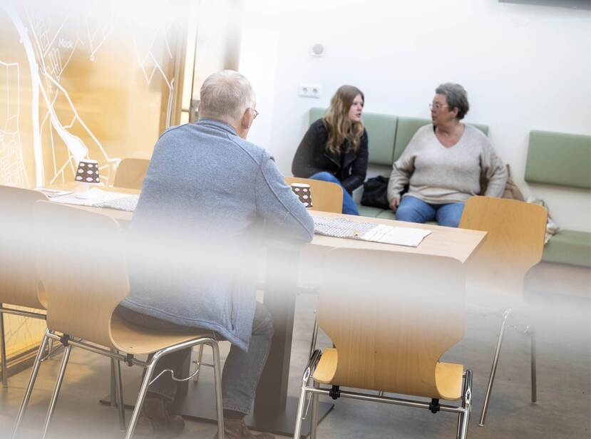 De foto toont een wachtkamer. Een oudere man met een blauw vest aan zit aan tafel op een houten stoel. De rug van de man is te zien op de foto. Achterin op de foto zitten een oudere en een jongere vrouw op een bank tegen de muur.