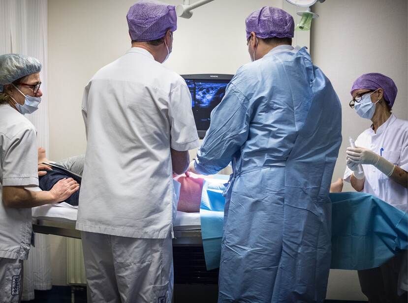 Op de foto staan 4 artsen om een operatietafel heen. Op de operatietafel ligt een patiënt onder een blauw zeil.