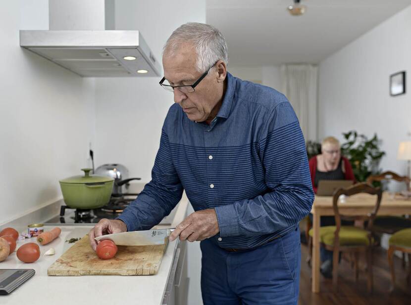 De foto toont een oudere man aan het aanrecht die een tomaat staat te snijden. Op de achtergrond zit een vrouw aan tafel achter een laptop.