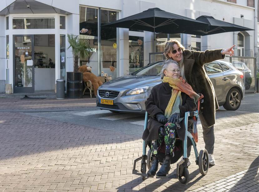 De foto toont een oudere vrouw in een rolstoel met achter haar een jongere vrouw. De vrouw die de rolstoel duwt wijst ergens naar met haar linkerhand. Ze staan op straat voor een horecagelegenheid.