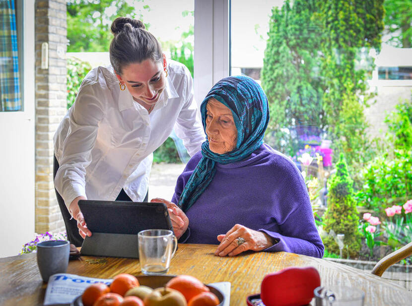 De foto toont een oudere vrouw met een paarse trui en een blauwe hoofddoek die aan tafel zit en op een tablet kijkt. Rechts naast haar staat een jonge vrouw in een witte blouse die meekijkt en uitleg geeft over iets dat op het scherm is te zien.