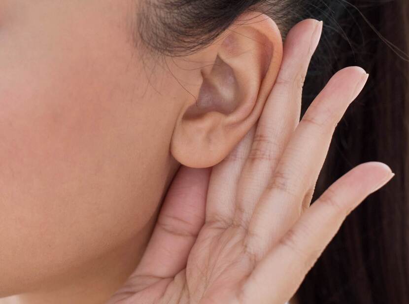 De foto toont een close-upbeeld van een oor, met een hand achter de oorschelp in een gebaar van 'wat hoor ik, wat zeg je?'.
