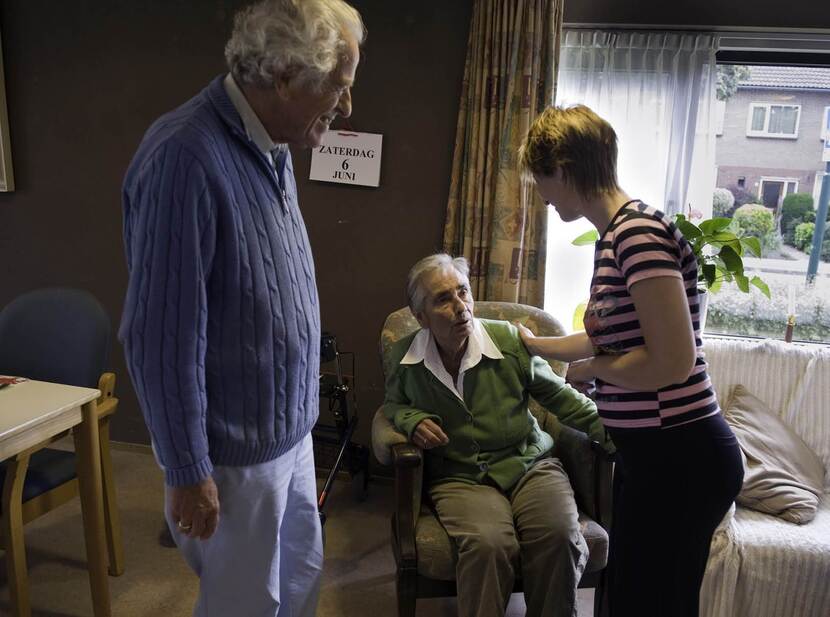 Het beeld toont een oudere vrouw zittend in een leunstoel. Haar schouder wordt aangeraakt door een jonge vrouw rechts van haar met een gestreept t-shirt aan. Links van naast de stoel staat een oudere man met een blauwe trui. Op de achtergrnond een rollator, een kalender waarop staat 'zatersdag 6 juni' en een raam met uitzicht op straat.